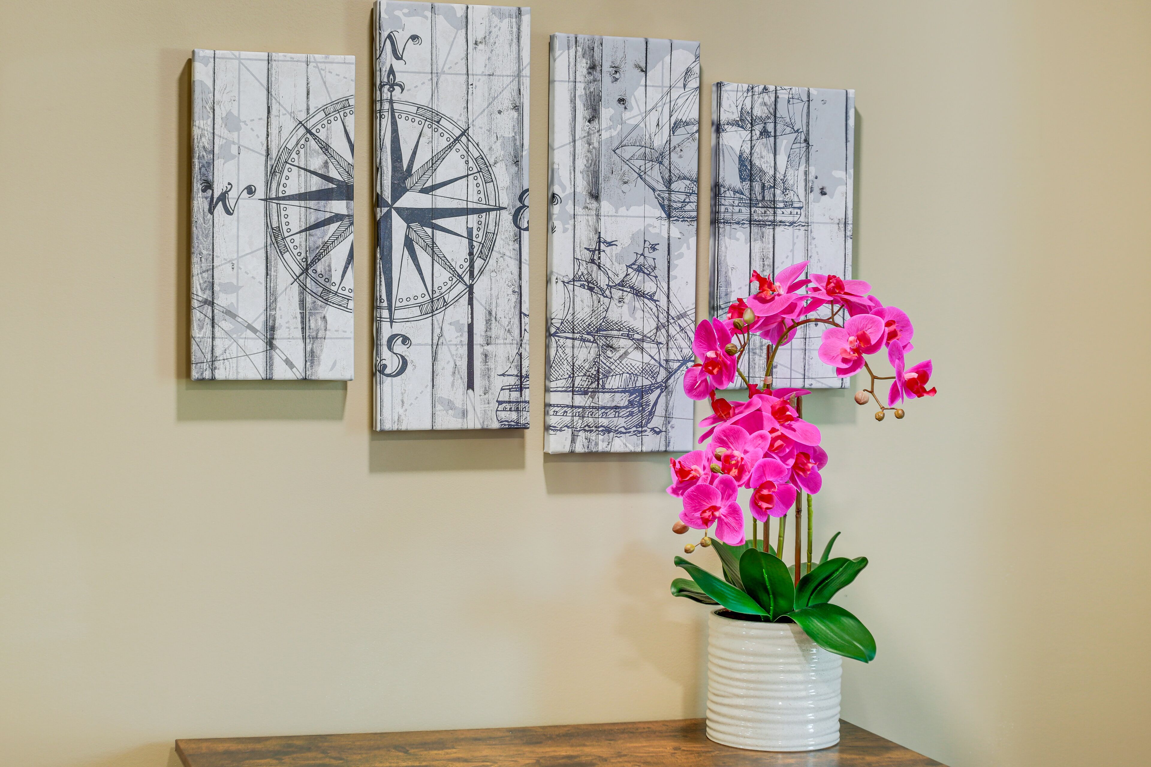 Una pared con tres cuadros enmarcados que muestran motivos marineros y un jarrón blanco con orquídeas rosadas.