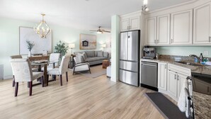 A modern kitchen with stainless steel appliances, a dining area with a glass table, and a living room with a gray sofa.