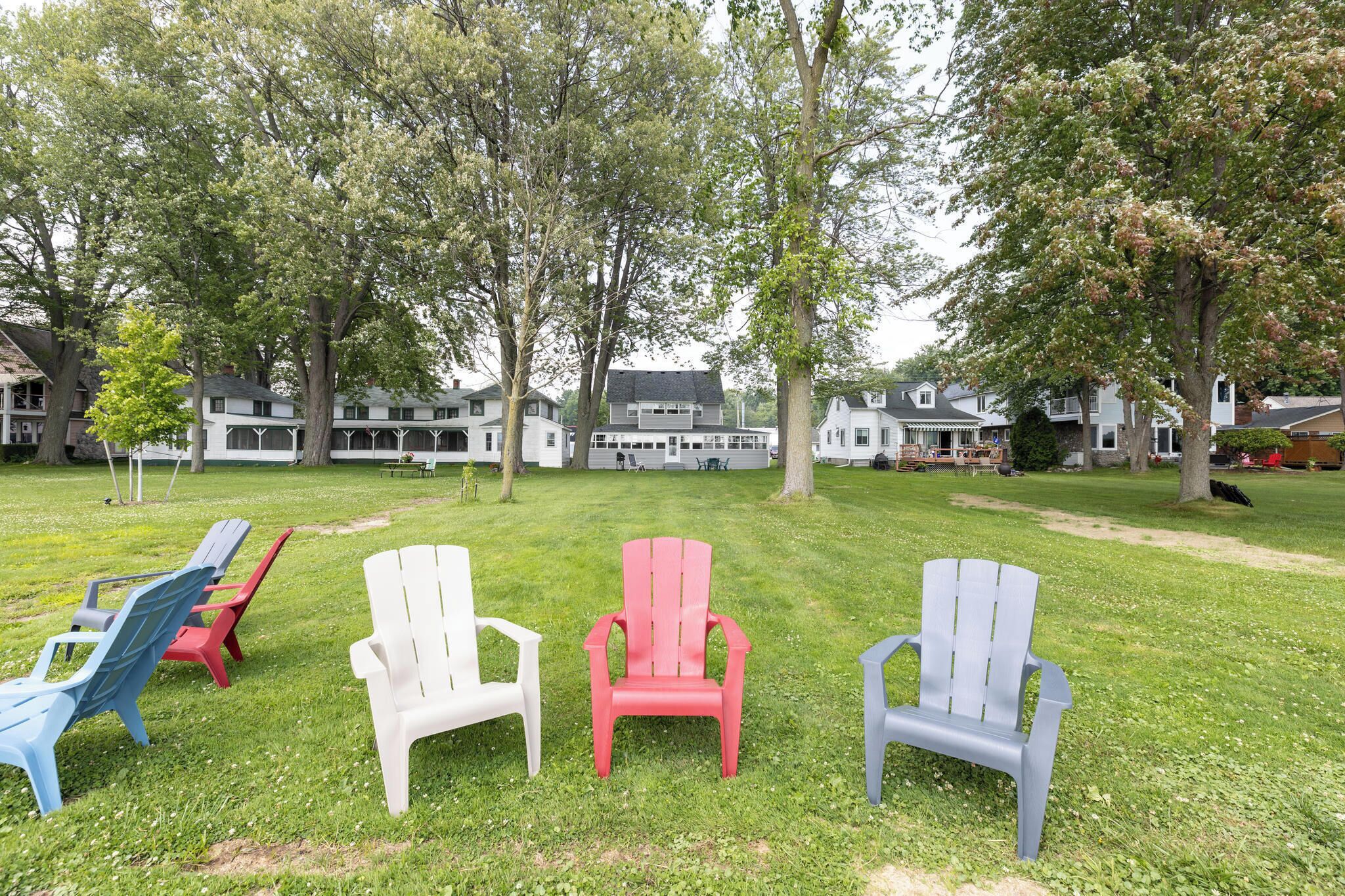 Een gazon met verschillende Adirondack-stoelen in uiteenlopende kleuren, een groot gebouw op de achtergrond en bomen.