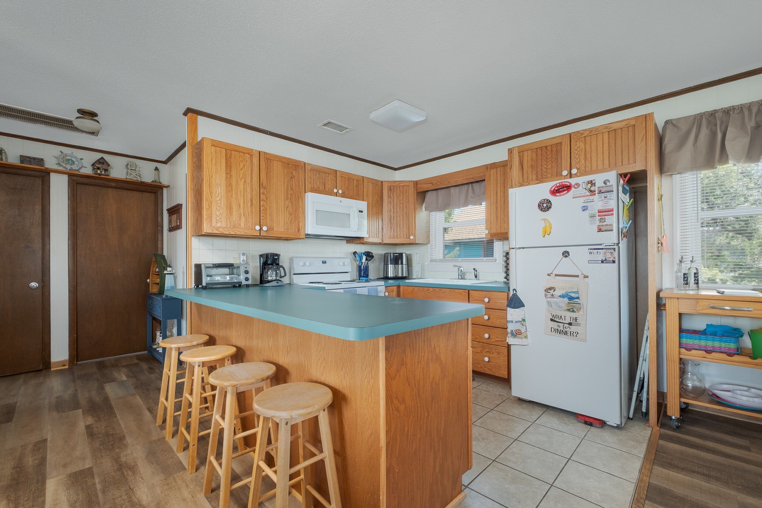 A kitchen with wooden cabinets, a white refrigerator, a microwave, and a blue countertop. There are three wooden stools and a window with curtains.
