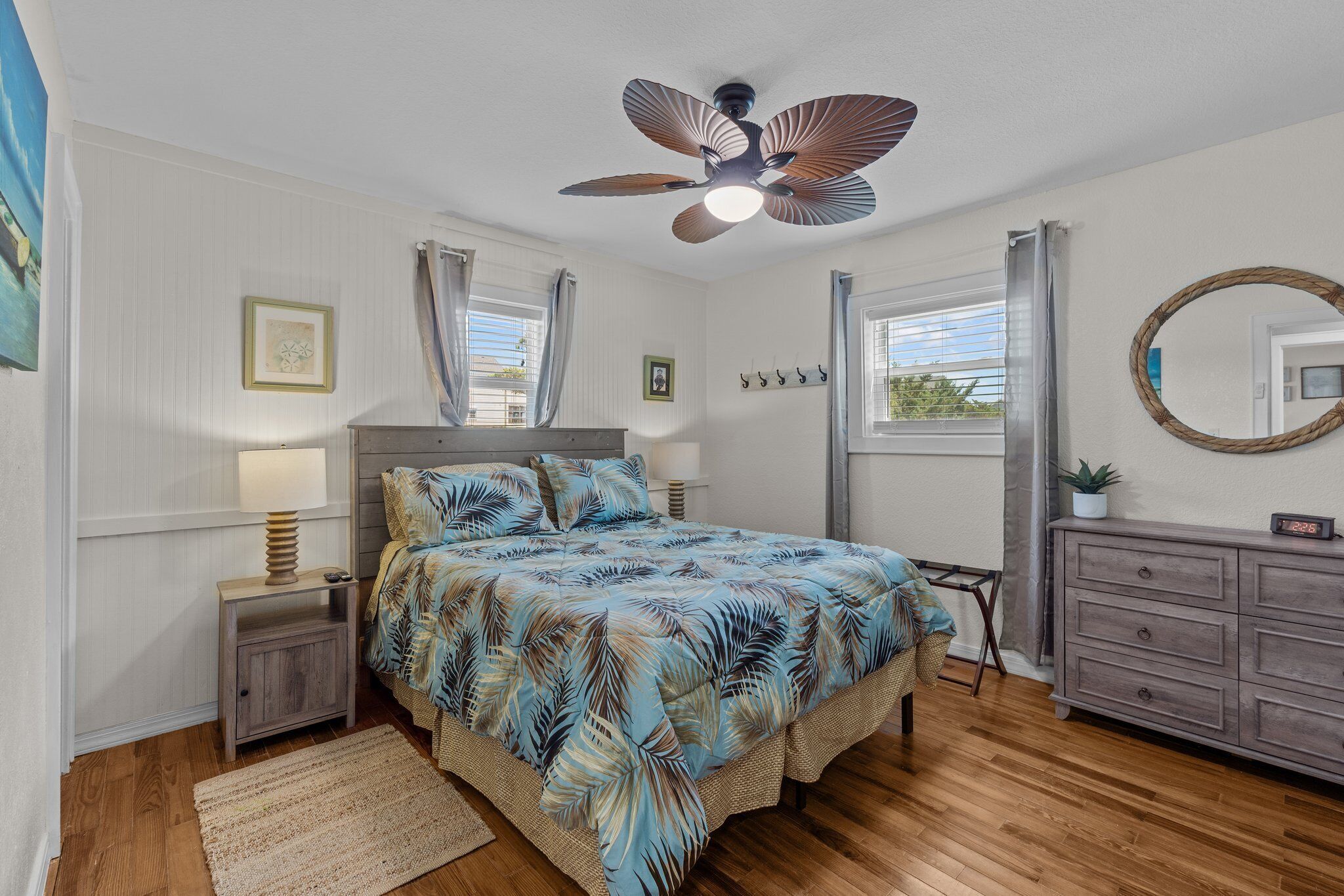 A bedroom with a bed, a ceiling fan, a nightstand, a dresser, a mirror, and a window with curtains.