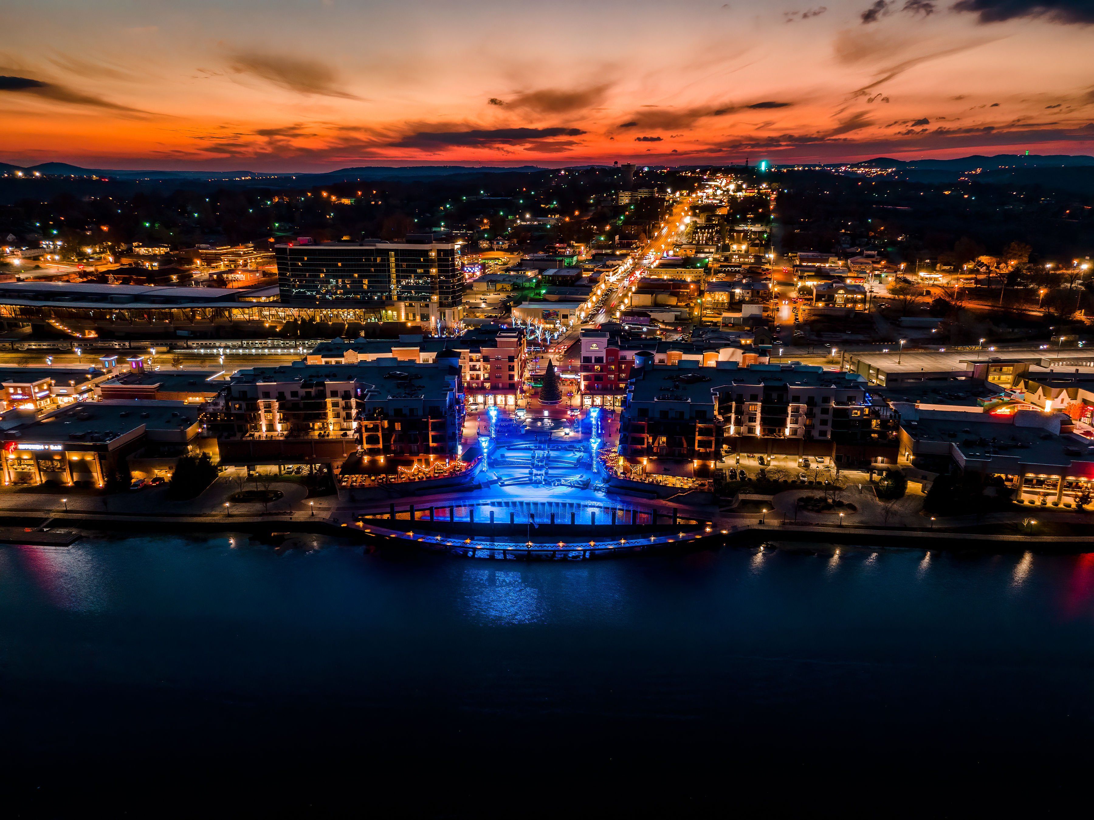 A waterfront cityscape at dusk with illuminated buildings and a prominent bridge structure.