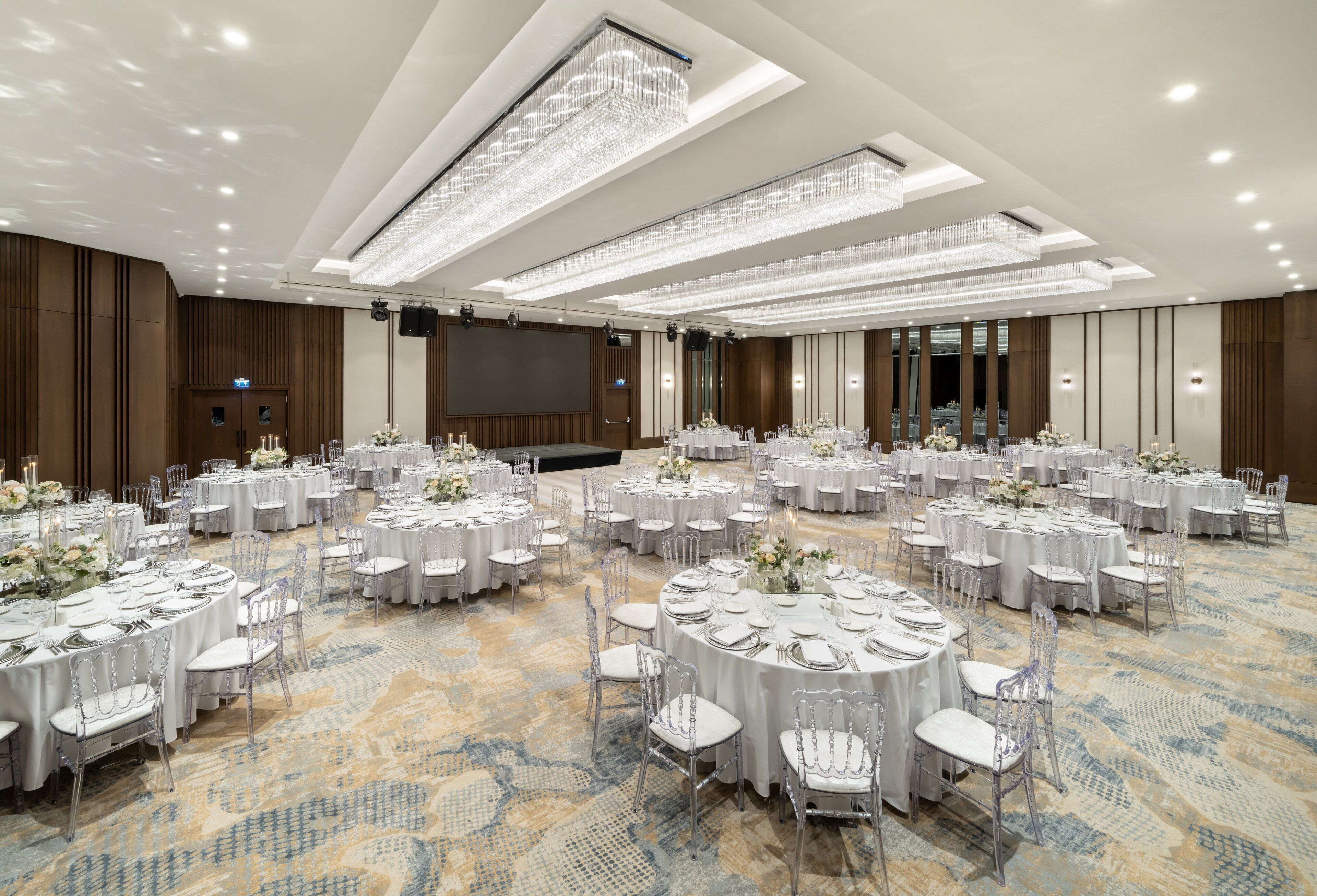 Um amplo salão de banquetes com mesas redondas preparadas para eventos, com carpete estampado e luminárias modernas.