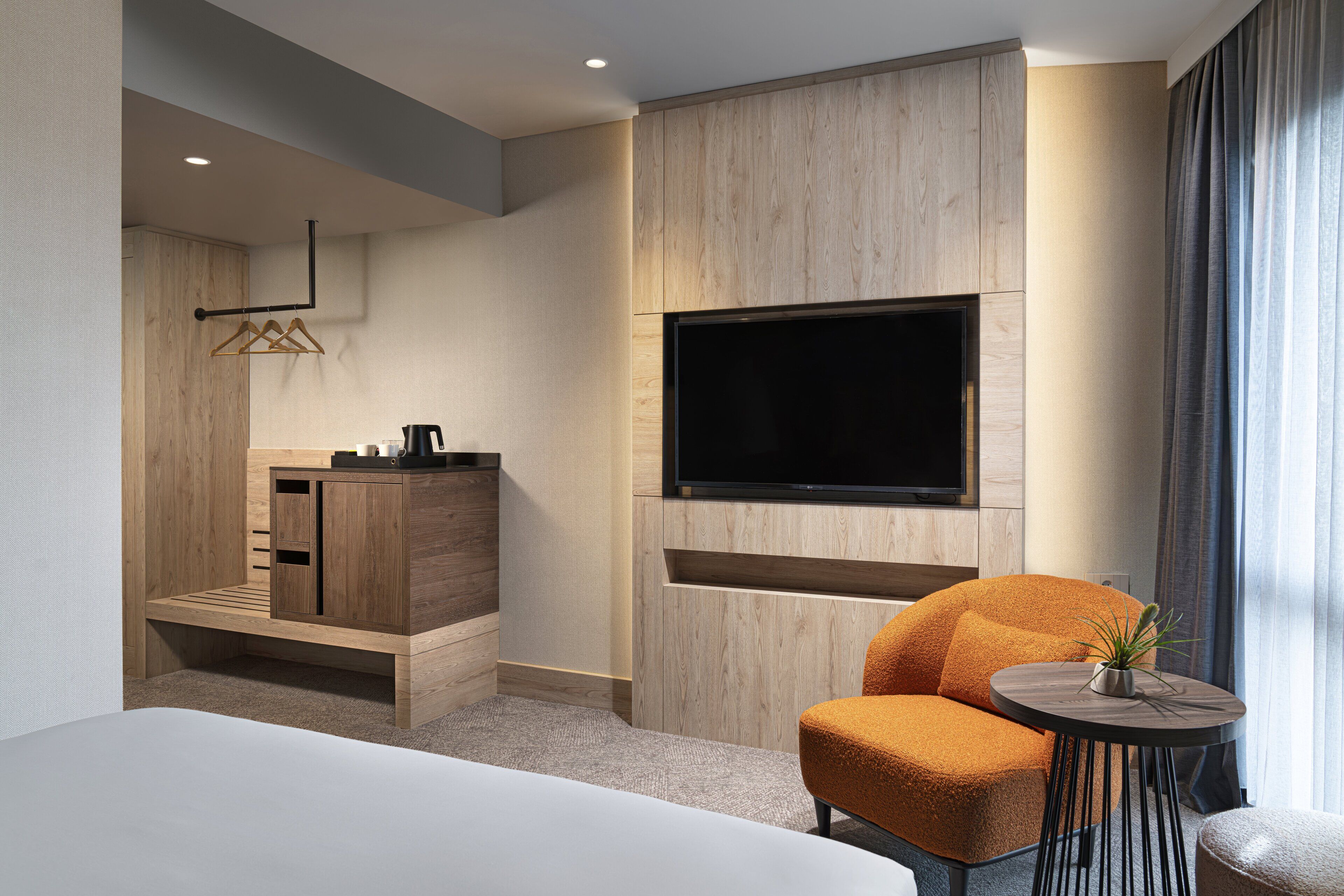 A modern hotel room with a large bed, a wooden cabinet, a flat-screen TV mounted on the wall, and a small round table with a plant.