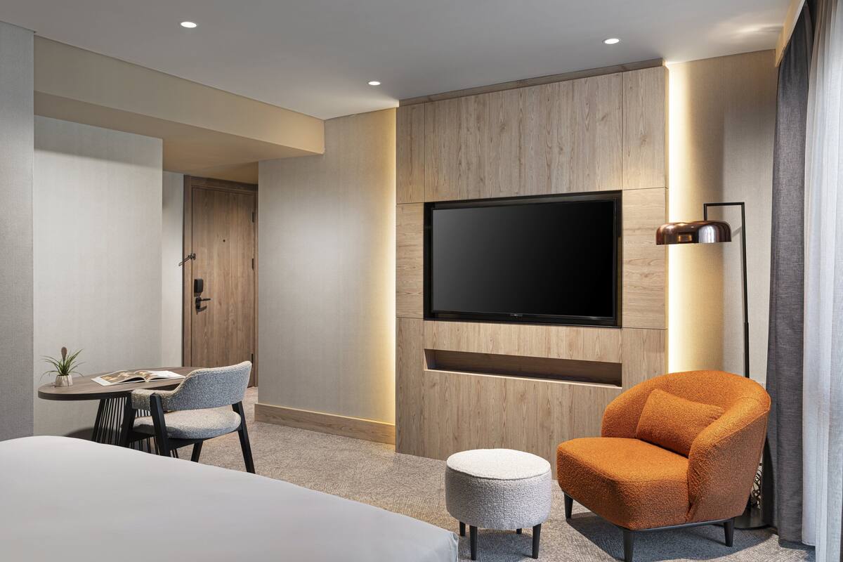 Une chambre d’hôtel moderne dotée d’un grand téléviseur à écran plat fixé au mur, d’une table de salle à manger ronde avec deux chaises, d’un petit pouf rond et d’un fauteuil orange.