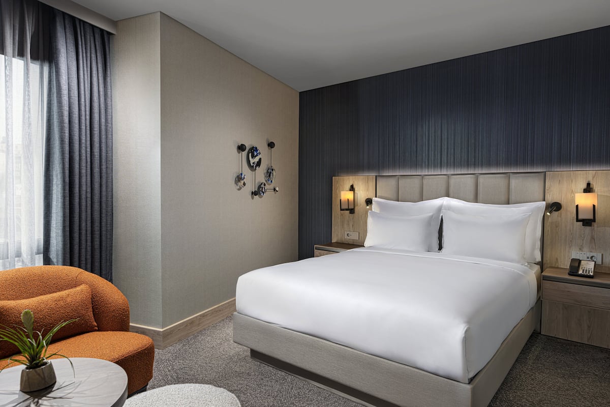 Une chambre d’hôtel moderne avec un grand lit, deux tables de chevet avec des lampes, un fauteuil rond orange et une petite table avec une plante.