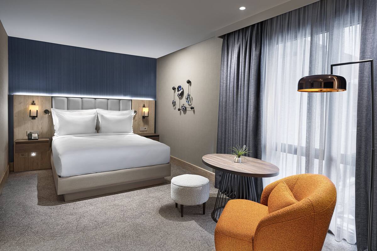 Une chambre d’hôtel moderne avec un grand lit, une table ronde agrémentée d’une plante, un fauteuil orange et une grande fenêtre avec des rideaux.