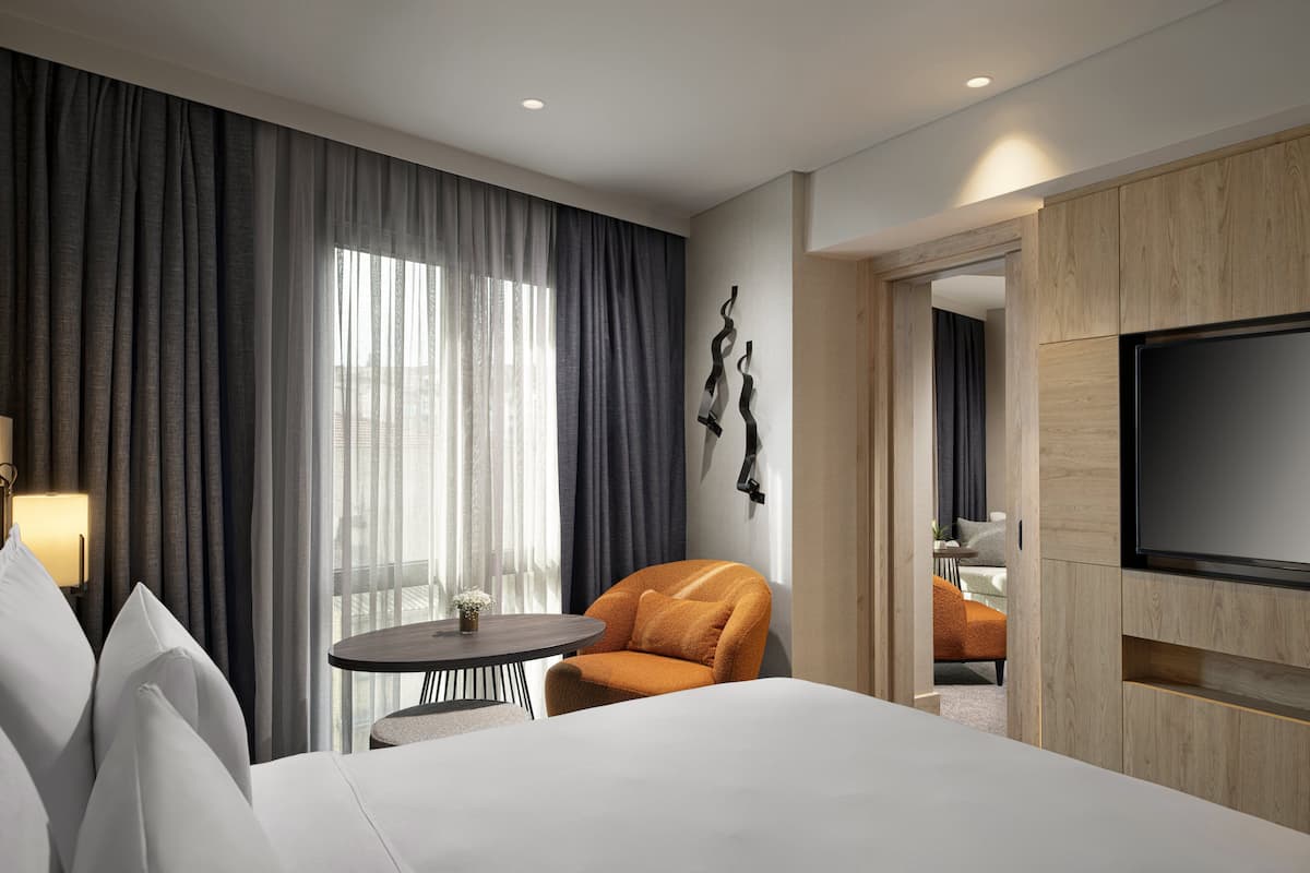 Une chambre d’hôtel moderne avec un grand lit, une table ronde, un fauteuil orange, une télévision et une vue sur la ville grâce à de grandes fenêtres.