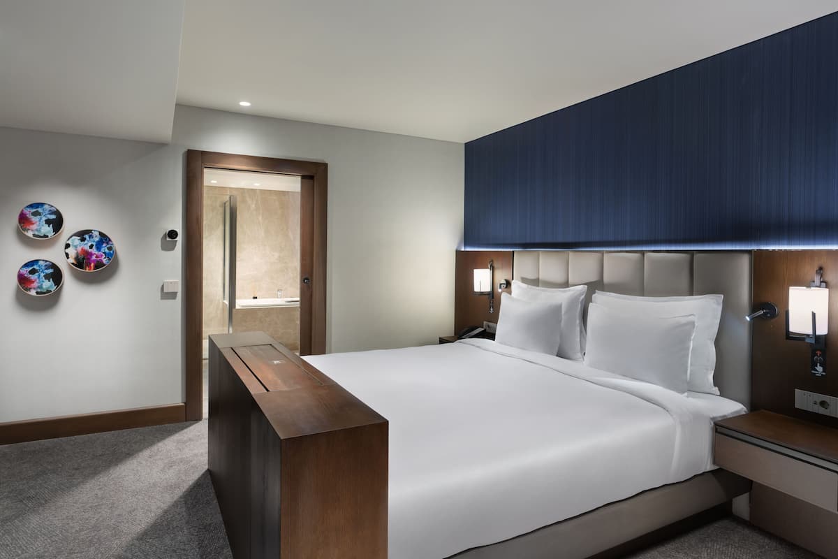 Une chambre d’hôtel moderne avec un grand lit, des tables de chevet, un banc en bois et des œuvres d’art décoratives au mur.