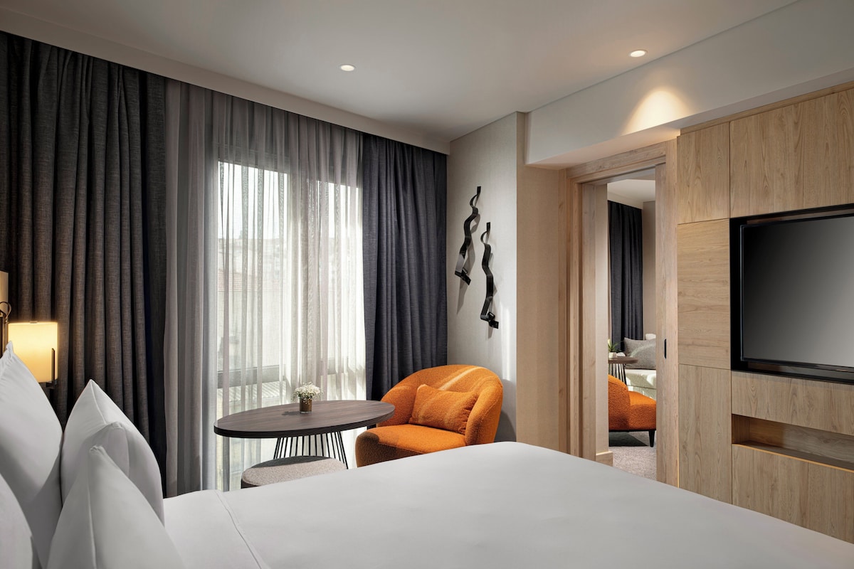 Een hotelkamer met een bed, een ronde tafel, een stoel, een televisie en uitzicht op buiten door grote ramen.