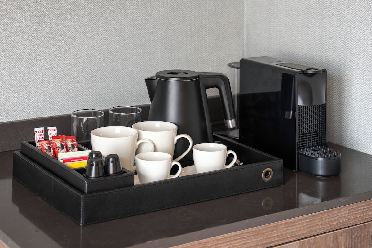 Een koffiehoek met een zwarte dienblad met mokken, een koffiezetapparaat en toevoegingen.