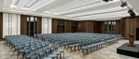 Een vergaderzaal met rijen stoelen, een spreekgestoelte en een plafond met lange lichtarmaturen.