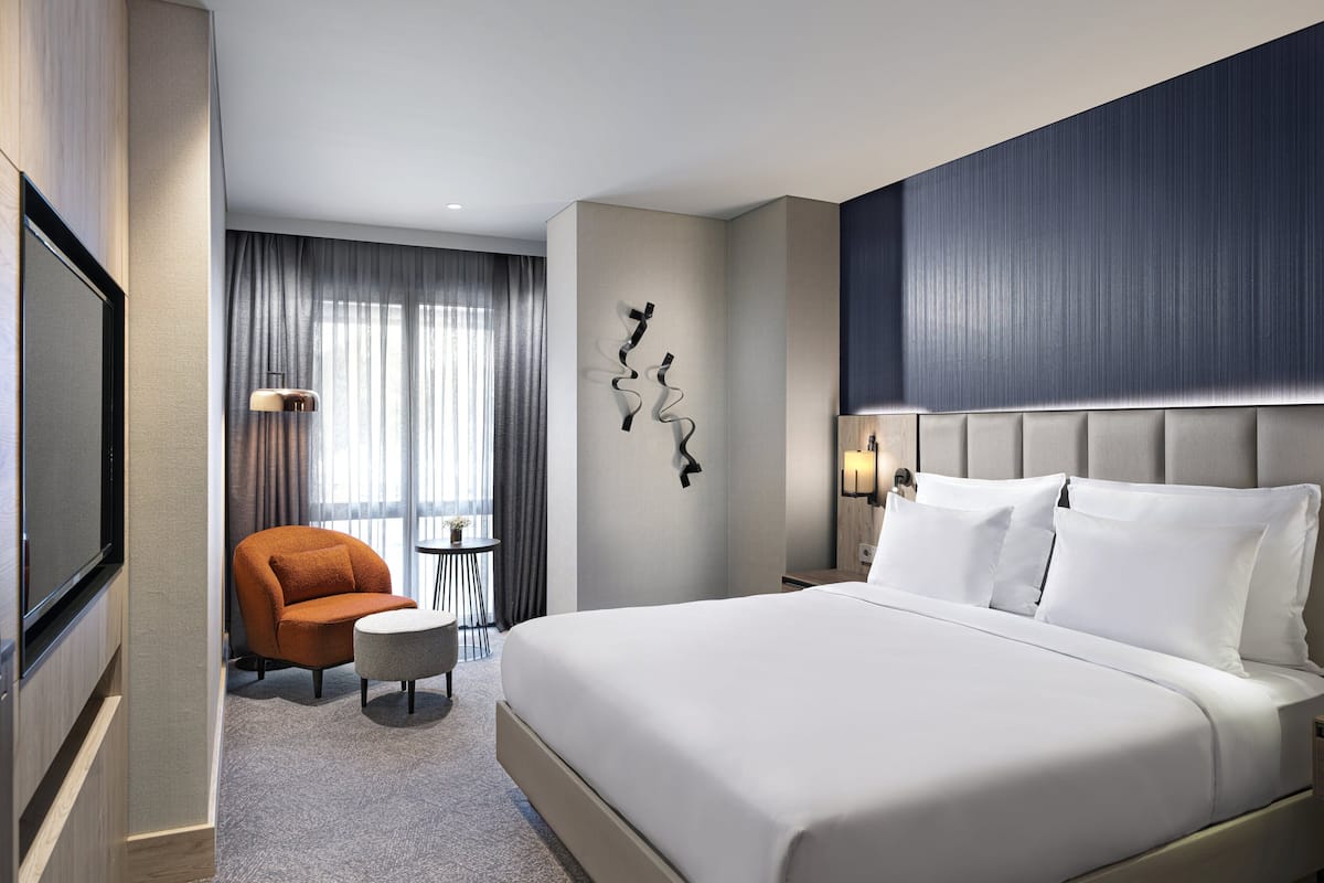 Une chambre d’hôtel moderne dotée d’un grand lit, d’un coin salon avec un fauteuil et un pouf, d’une télévision à écran plat et d’un élément décoratif mural.