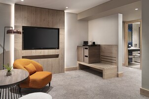 A modern living room with a flat-screen TV mounted on a wooden wall, a comfortable orange armchair, a small round table with a plant, and a bench with storage cabinets.