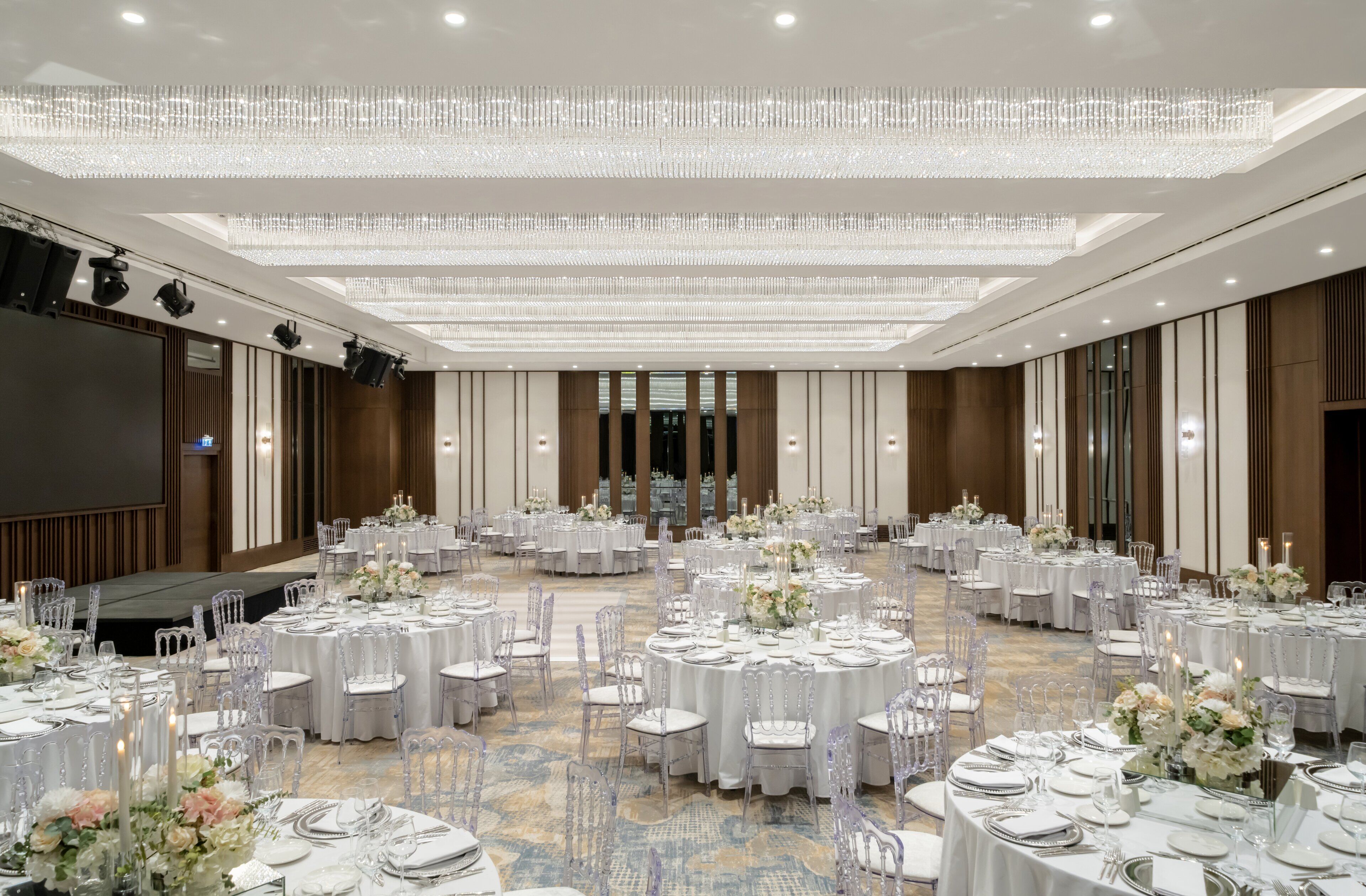 A large banquet hall with round tables set for events, featuring chandeliers and a stage area.