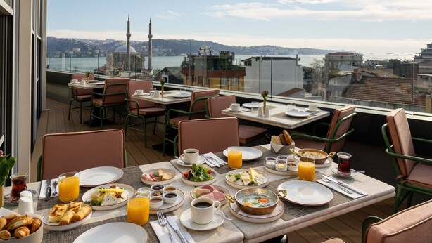 Un área de comedor en la azotea con mesas dispuestas para el desayuno, con vistas a un paisaje urbano que incluye edificios y una masa de agua.