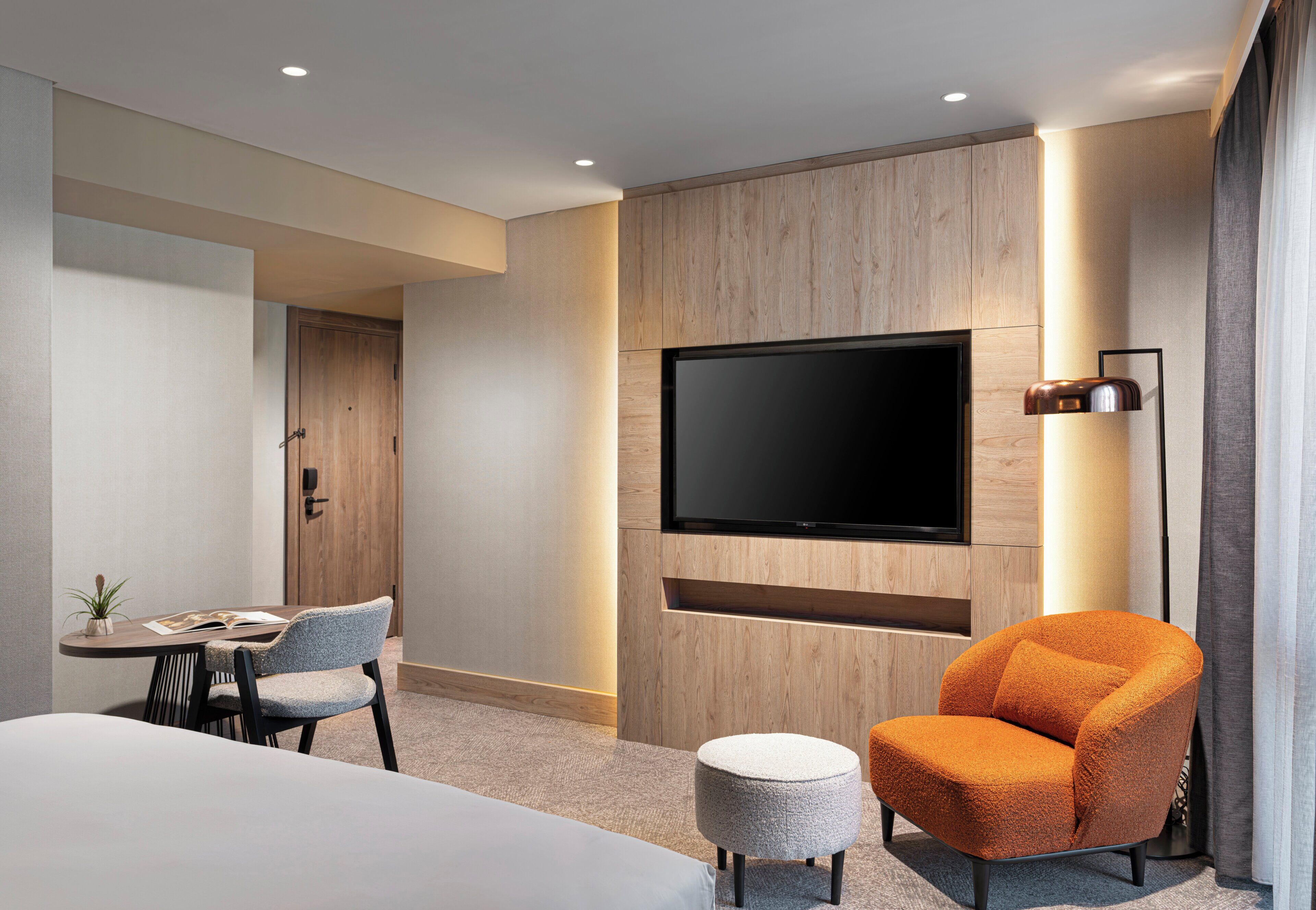 Ein modernes Hotelzimmer mit einem Wand-Fernseher, einem runden orangefarbenen Sessel, einem kleinen runden Hocker und einem Essbereich mit Tisch und Stühlen.