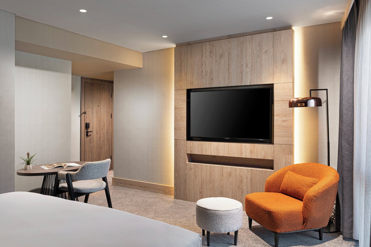 Een moderne hotelkamer met een flatscreen televisie aan de muur, een ronde oranje fauteuil, een klein rond voetenbankje en een zithoek met een tafel en stoelen.