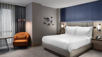 A hotel room with a large bed, a nightstand with a phone, a chair, a table, and a wall-mounted light fixture.