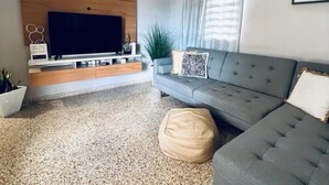 Ein modernes Wohnzimmer mit grauem Sofa, einem Flachbildfernseher an einer Holzwand, einer Topfpflanze und einem strukturierten Teppich.