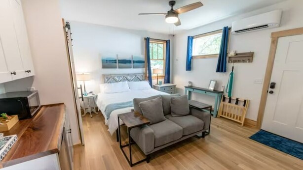 Ein modernes Schlafzimmer mit einem Bett, einem grauen Sofa, Holzboden und Deckenventilator.
