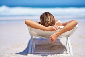 A person reclining in a white chair on a sandy beach with the ocean in the background. - Les Dunes 1 (Plonévez-Porzay)