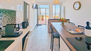 Una cocina moderna con cafetera, un comedor con mesa y sillas, y vista al mar a través de amplios ventanales.