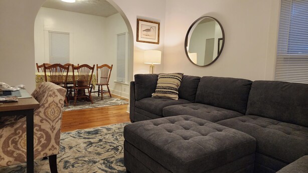 A living room with a sectional sofa, a dining table with chairs, a patterned rug, and a round mirror on the wall. - Great for working professionals: medical, construction, etc. LTE, or Mid-term! (Grand Rapids)