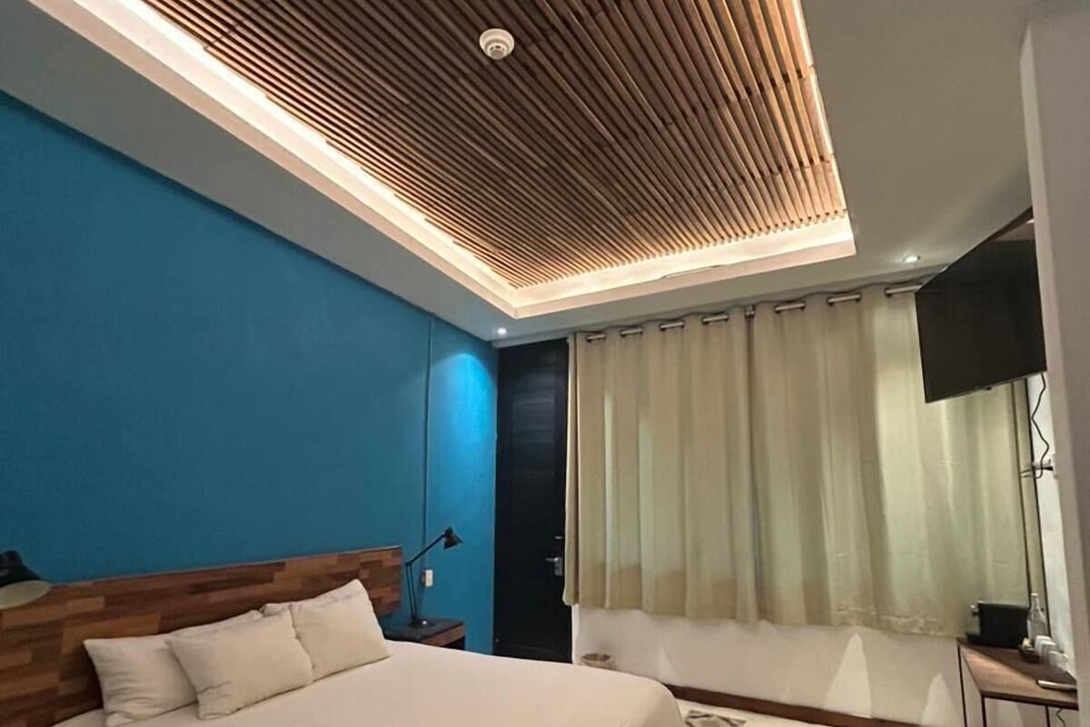 Quarto moderno com teto de madeira, uma cama grande, mesa de cabeceira, televisão e janela com cortinas.