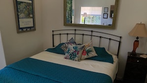 A neatly made bed with teal bedding and floral pillows, a bedside table with a lamp, and a framed picture on the wall.