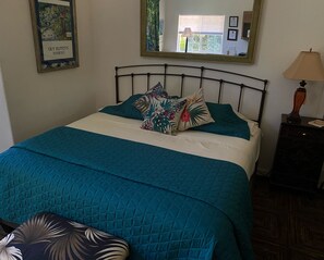 Un lit bien fait, avec une literie de couleur turquoise et des coussins à motifs floraux, une table de chevet avec une lampe et un tableau encadré au mur.
