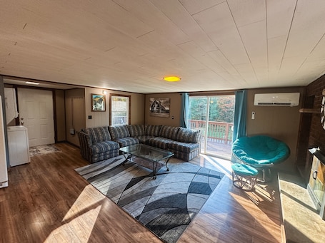A living room with a sectional sofa, a glass coffee table, and a teal bean bag chair. There is a sliding glass door leading to a deck.