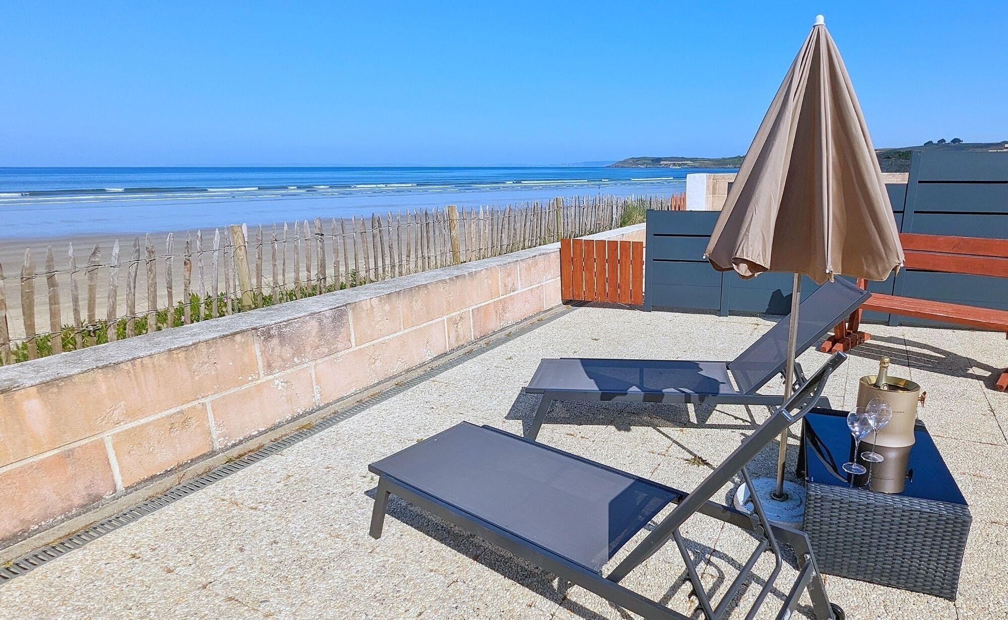 Una terrazza sul tetto con vista sulla spiaggia, una recinzione in legno e un tavolo con una bottiglia e dei bicchieri.