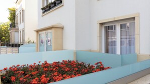 A building with a light blue exterior, a flower bed with red flowers, and a window with a white curtain. - Studio with Balcony, Business Studio, 1 Twin Bed  (LUXEMBOURG)