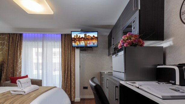 A hotel room with a bed, a desk with a computer, a television displaying a cityscape, and a clock on the wall. - Studio with Balcony, Business Studio, 1 Twin Bed  (LUXEMBOURG)