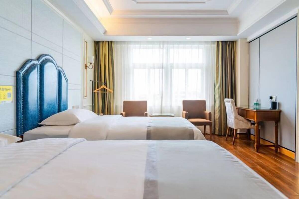 Habitación de hotel con dos camas, un escritorio, sillas y un ventanal con cortinas.