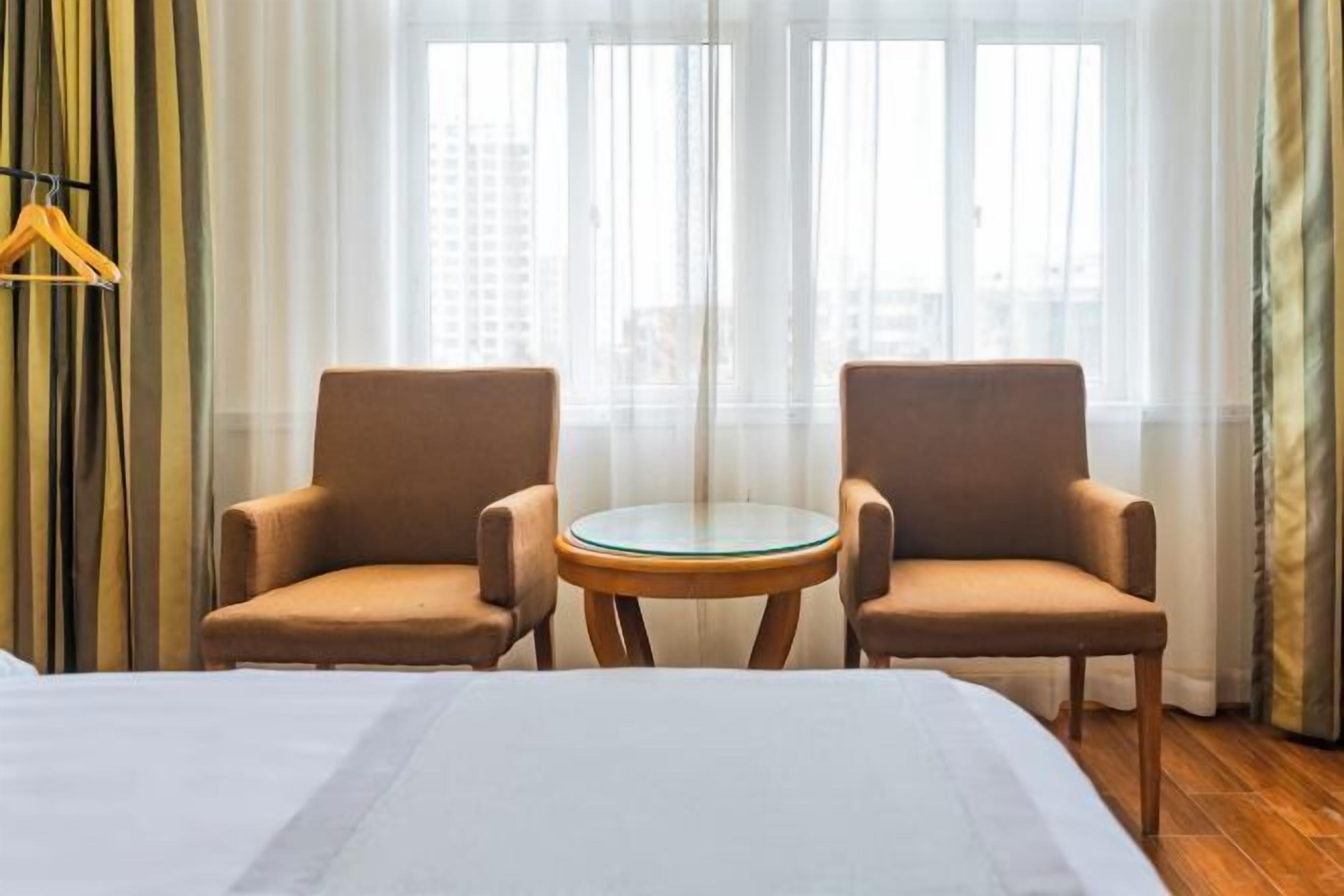 A hotel room with a bed, two armchairs, a small round table, and a view of buildings through large windows.