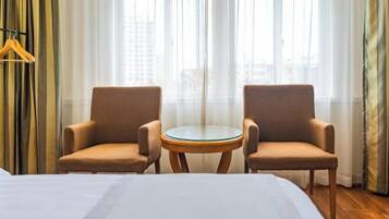 A hotel room with a bed, two armchairs, a small round table, and a view of buildings through large windows.