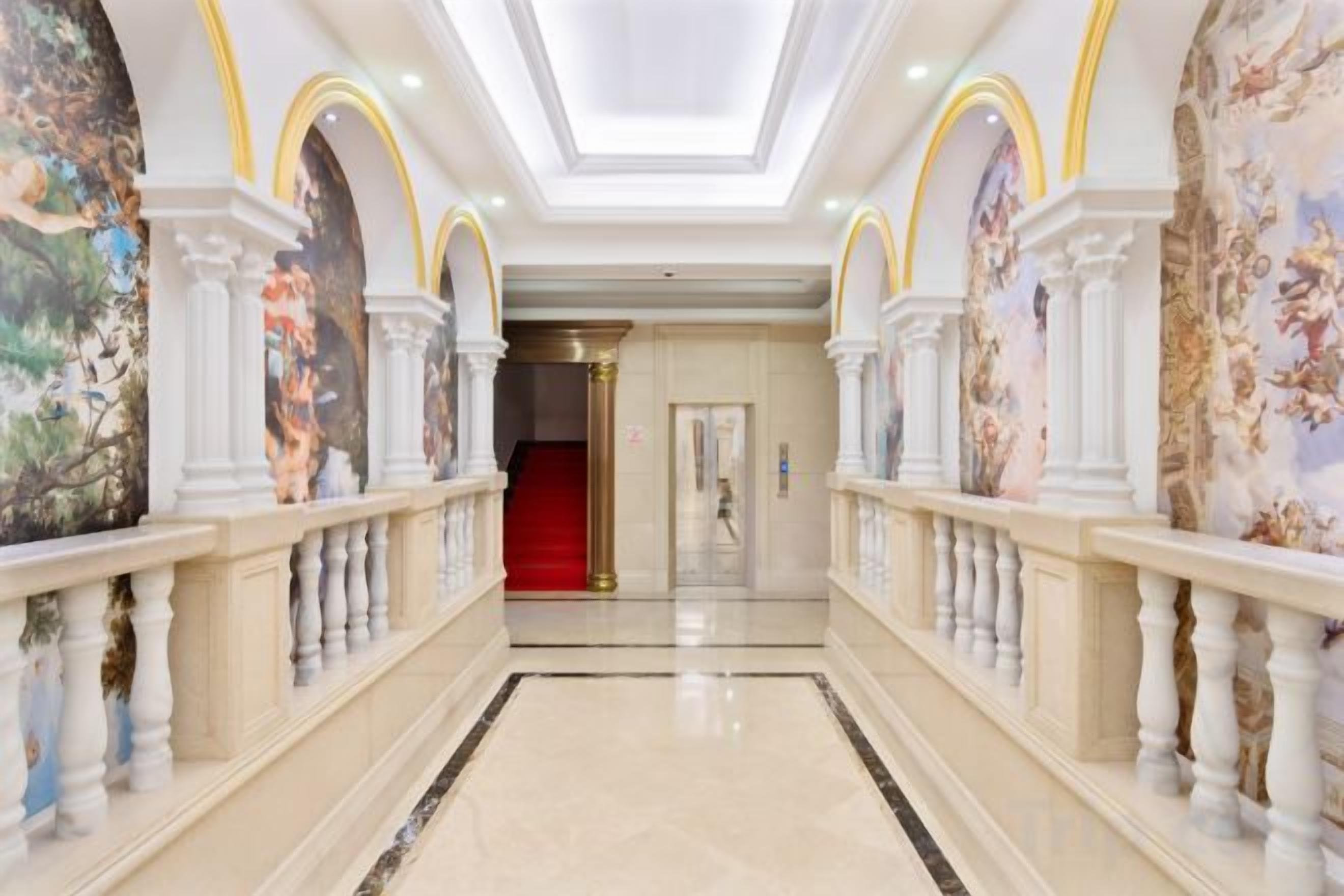 A grand hallway with a marble floor, white columns, and a red carpet leading to a door.