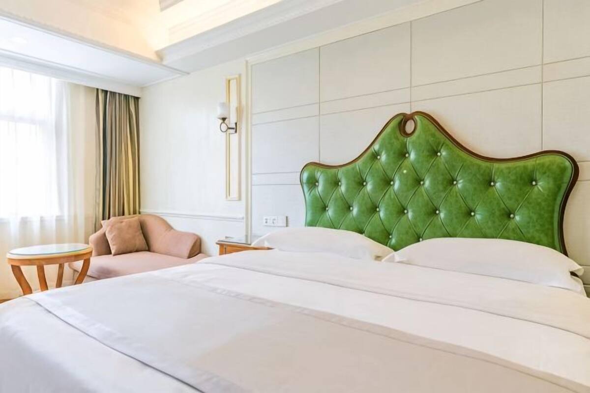 Una habitación de hotel con una cama grande, un cabecero tapizado en verde, una pequeña mesa redonda y una silla.
