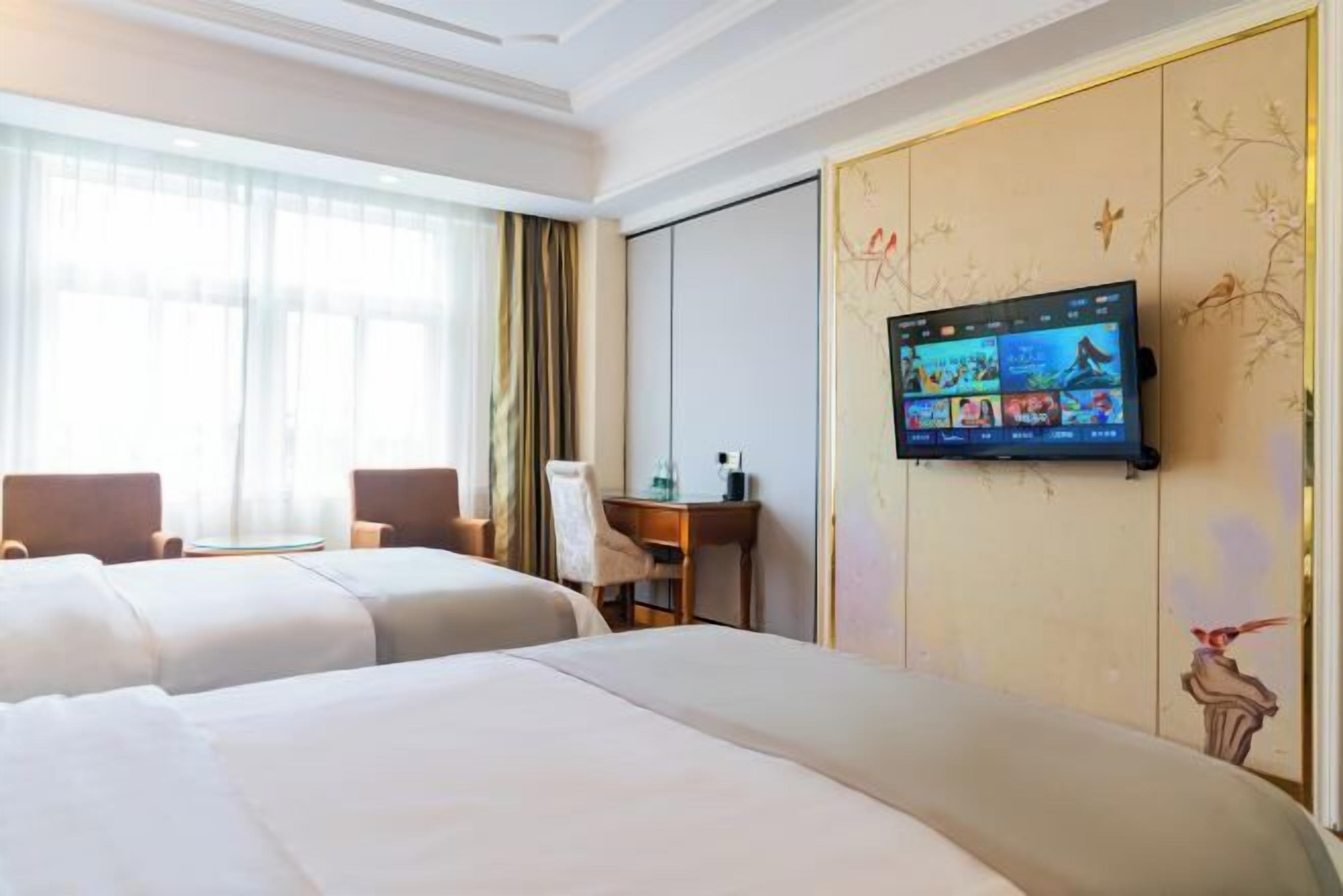 A hotel room with two beds, a desk, and a TV mounted on the wall displaying various channels.