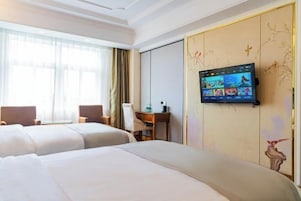 A hotel room with two beds, a desk, and a TV mounted on the wall displaying various channels.