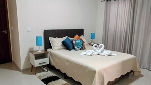 Un letto rifatto con una coperta bianca e due cuscini, una lampada blu su un comodino, un asciugamano decorativo a forma di cigno sul letto e un tappeto a righe sul pavimento.