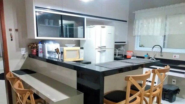 Una cucina moderna con un piano di lavoro nero, mobili bianchi, un forno a microonde e un frigorifero. Due sedie di legno con cuscini neri sono presenti al bancone.