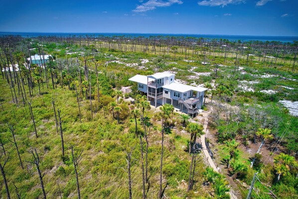 A large, modern house with multiple levels and a balcony, surrounded by a natural landscape with sparse vegetation and a view of the ocean.