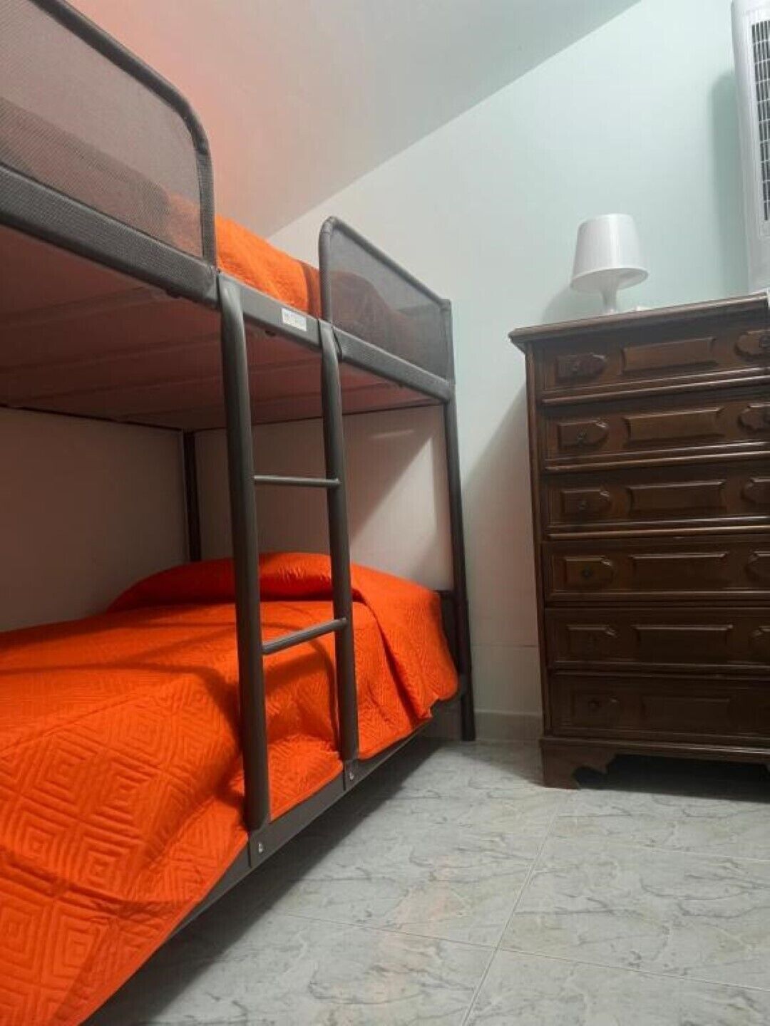 Una cama individual en literas con ropa de cama naranja, un aparador de madera y una lámpara blanca.