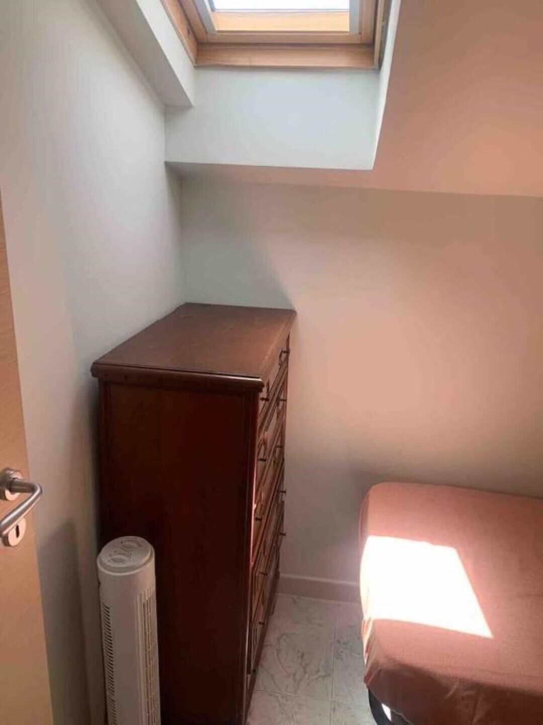 A small room with a skylight, a wooden dresser, a radiator, and a bed with a light-colored bedspread.