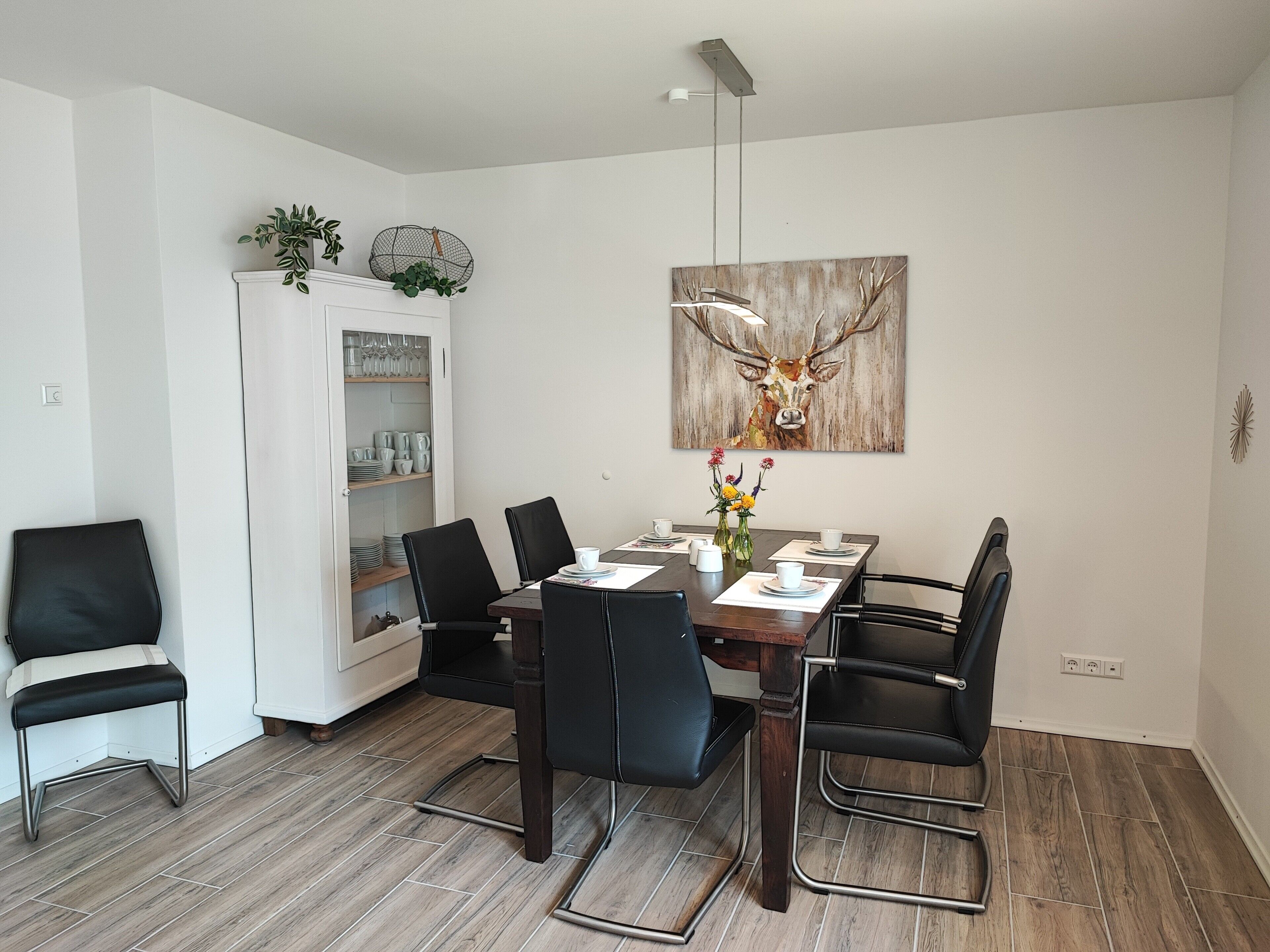 A dining area with a wooden table, black chairs, a cabinet with dishes, and a deer painting on the wall.