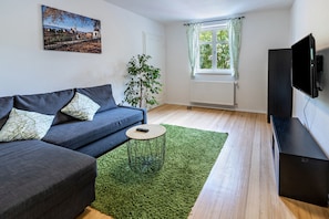 A modern living room with a sofa, a small round table, a green rug, a plant, a window with curtains, and a TV.