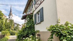 A narrow street with a church steeple, a building with green shutters, and lush greenery.